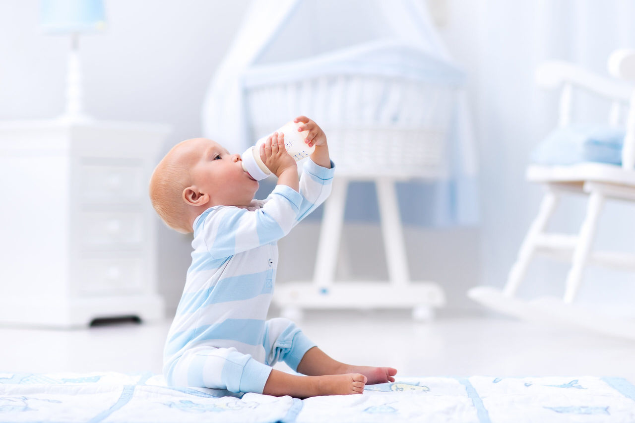 Adorable baby boy playing on a blue floor mat and drinking milk from a bottle in a white sunny nursery with rocking chair and bassinet. Bedroom interior with infant crib. Formula drink for infant.; Shutterstock ID 481192738; purchase_order: DNC thumbnails ; job: Allergy papers; client: ; other: 